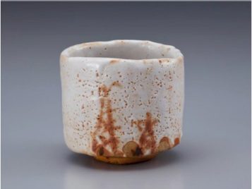 The image shows a short tea bowl with vertical walls and an uneven rim. The glaze is white with small holes or pits across the surface. In places along the top lip and across the sides, orange-brown color - almost the color of rust - seems to push through the thicker white glaze. Coming from the bottom, a few orange-brown vertical lines reach toward the top, going halfway or one third of the way up the side. Smaller horizontal lines cross these, creating a faded image of bamboo growing. Along the bottom of the tea bowl, the glazes and colors end, leaving what looks like the raw clay exposed. The clay is tan except in places where the orange-brown glaze touches it, where it is dark brown or almost black.