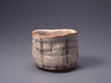 The image shows a small tea bowl with vertical sides and that is glazed in an off-white color. The top edge and sides are slightly uneven and include small marks, indentations, and places where it looks like the glaze has not fully covered the clay. The lip of the tea bowl seems to be stained with a rust-colored material that is lighter and darker around the top. On the walls of the tea bowl, simple and rough lines have been painted vertically and horizontally to suggest a fence. More of the rust color occurs around the bottom of the tea bowl, but not in any obvious shape or pattern.