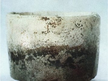 This image shows a small tea bowl with rough, vertical sides. The colors create a horizon line across the sides, with a largely white or cream upper half and a bottom half that is mottled brown, tan, and black. The edges are all slightly uneven and the colors seem to randomly bleed into each other. This is the same tea bowl pictured in the lower left corner of the previous image.