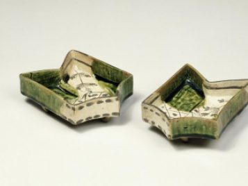 This image shows two small dishes shaped like arrows. The dishes have fully flat bottoms and vertical walls, and the hint of stubby feet holding them up. They are glazed with a white color with transparent green areas. The areas with only white are also decorated with black or dark brown color, making dashes, lines, dots, or the patterns of tree branches. It has the effect of creating visual busy-ness.