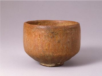 This tea bowl has vertical walls that turn into a gentle sloping bottom, and a small lip of a foot at the very bottom. It is not immediately obvious if the bowl is glazed or if it is naked clay. If there is a glaze, it seems very natural. The tone is red and orange, with small places of white and brown. Although the tea bowl is even, its surface is not fully smooth and it includes small differences in both the color and texture.