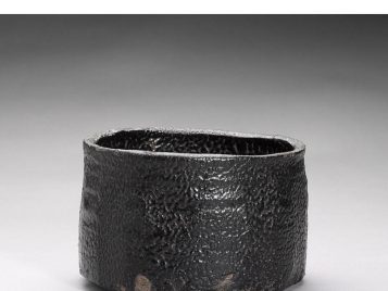 The image shows a small tea bowl with vertical sides and that is glazed in black and shiny. The tea bowl seems to have a texture that seems almost like leather or lizard skin. The top edge and sides are slightly uneven but thick. The edges of the tea bowl do not taper much at the top, but stay thick. It seems like the walls of the tea bowl extend almost to the very bottom, with no "foot" underneath it, but there is a very small shadow, suggesting something is on the very bottom of the bowl but not visible from this angle. 