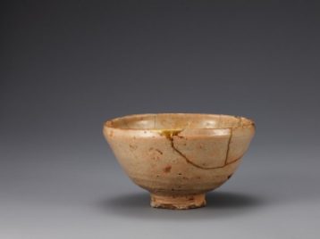 This tea bowl is tan, light brown, and peach in tone. The side is gently curved to the bottom. This bowl has been restored following breakage into ten pieces. The repairs are made in gold and are shiny compared to the rest of the bowl. Its rim curves inwards. It is made with stoneware clay. There are five spur marks on the inner base, and the glaze on the rim of its foot has been wiped away. It is coated with opaque white glaze, on which have formed many crackles and pinholes. The color of glaze is not even throughout the surface. 