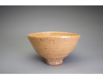 This tea bowl has sloping sides and seems to be the color of clay. It is a peachy tan color, with brown or black spiderweb shapes across the surface. Rather than being painted on, those shapes seem to reflect cracks in the glaze or other imperfections in the clay. The tea bowl seems to have been glazed but with a glaze that is very thin. The bowl's surface is slightly shiny overall, but at the very foot, the white glaze has built up into larger puddles. Clusters of the white glaze is only obvious at the very bottom of the bowl, as if it had pooled there.