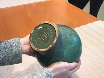 This image is a photograph of a person's hands holding a green vase, which is flipped upside down so we can see the very bottom. The vase itself is glazed in shades of green that look very much like the color of bronze when it oxidizes. The very bottom of the vase is flat and has the stylized signature of the artist. His initials - CFB - are carved into the clay on top of each other so that they resemble a Chinese character. Below this, we see 19 / 15, the year the piece was made.