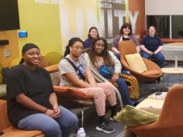 A diverse group of students are arranged in a conversational circle in a lounge at Alice Lloyd Hall, with a visiting artist. The lounge is decorated with comfortable orange chairs, sofas and tables. And there is a mural on the wall behind them that spells out UNITY, done in browns, greens and oranges.