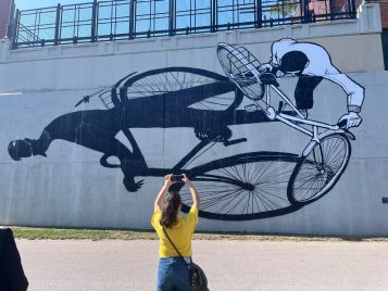A student with a long brown ponytail and wearing a bright yellow t-shirt and jeans is pictured in the foreground facing away from the camera. They are taking a photo of a large black and white mural of a cyclist and his shadow on the side of a concrete wall.