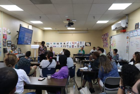 A group of people seated in a classroom listening to a presentation. The room has several tables, a whiteboard, and various posters on the walls.