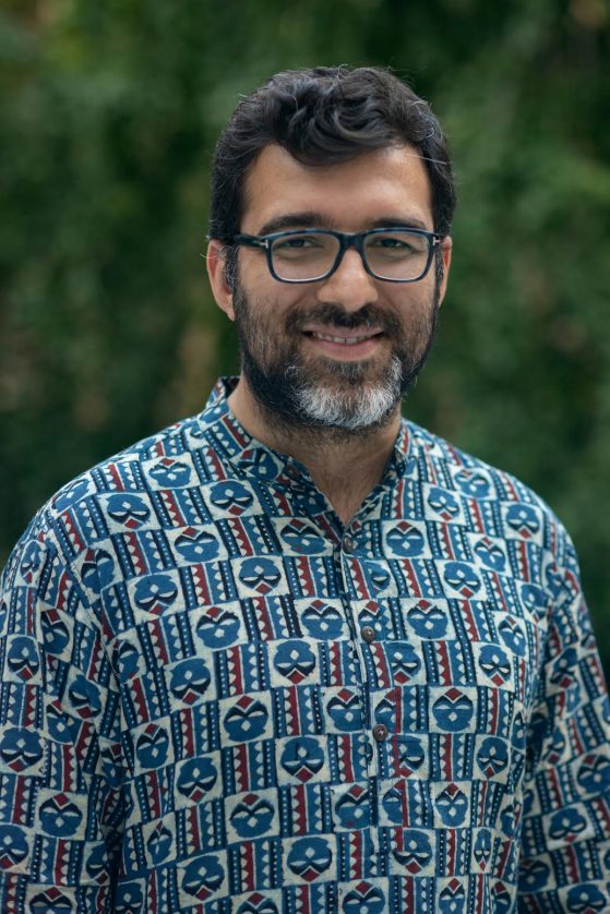An individual with a beard and dark hair smiles directly at the camera while wearing a blue patterened shirt