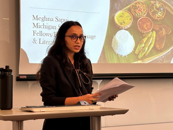 Individual with long hair wearing a black jacket, speaking at a podium in front of a powerpoint title slide. 