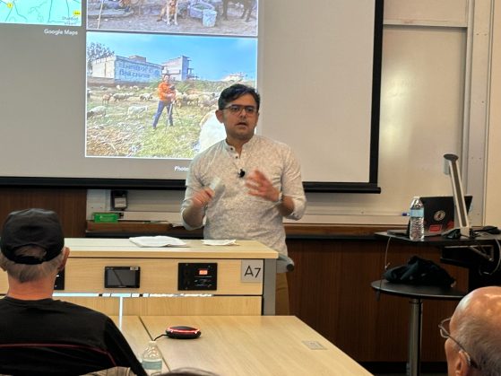 Harjant Gill is at the front of the room by a podium presenting in front of a projector slide