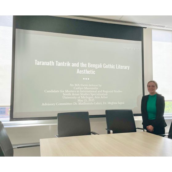 Caitlyn is standing in front of a projecter screen displaying the title of her thesis on a green background. 