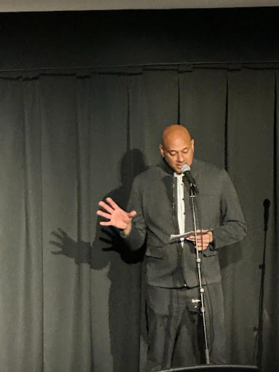 A man wearing all black stands on a stage in front of a microphone reading off an index card