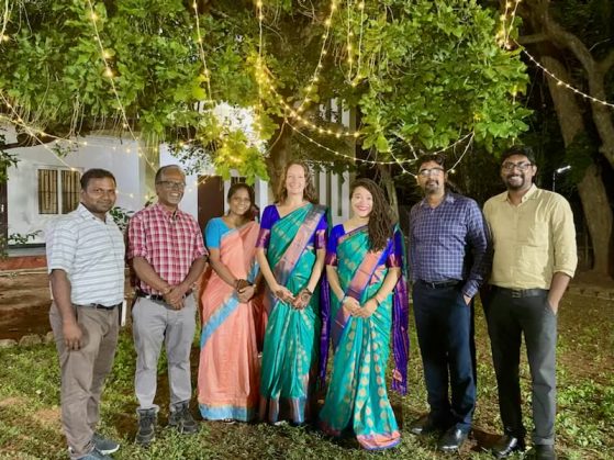 Four men and three women pose together in the evening in front of a white home and tree with string lights