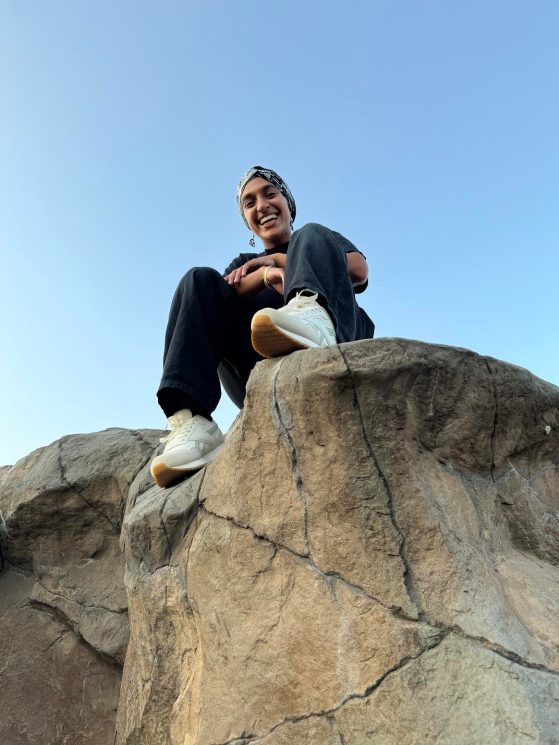 Image is looking up at an individual sitting on a large rock smiling at the camera and wearing tennis shows, dark clothing, and a head wrap