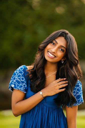 A person with long, wavy brown hair, wearing a blue shirt is standing in a green field with her hand laid across her chest.