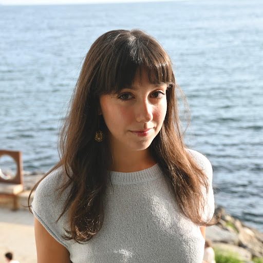 A person with long, wavy brown hair is outdoors, wearing a short-sleeved light top. In the background, there's a body of water and a bench on a sandy or rocky shore, conveying a calm, sunny setting.