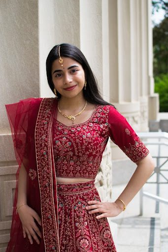A person is outdoors wearing a red traditional outfit with gold embroidery, including a half-sleeve blouse, long skirt, and a sheer scarf. They accessorize with a necklace, bangles, and decorative hairpiece. The background shows a stone building with columns and greenery.
