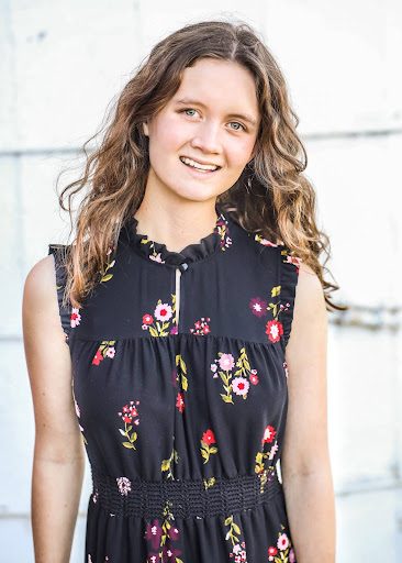 A person with shoulder-length, wavy brown hair is wearing a sleeveless black dress adorned with colorful floral patterns. The background features a light-colored wall.