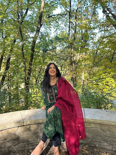 A person is outdoors wearing a green patterned outfit with a red draped scarf. They are sitting on a low stone wall in a wooded area with lush green trees and sunlight filtering through.