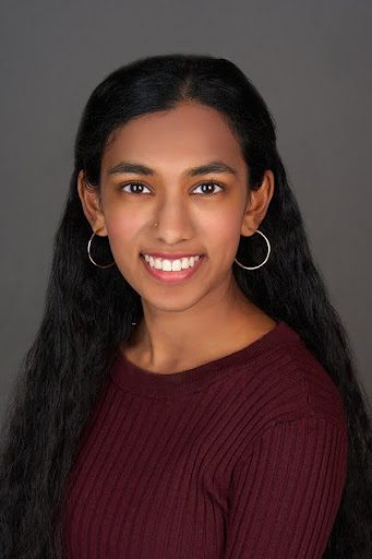 A person with long, straight black hair is wearing a maroon ribbed top and large hoop earrings. The background is a plain gray color.