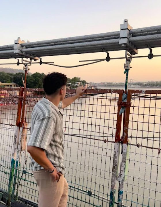 An individual wearing a green striped shirt is standing on a bridgelooking off into the sunset