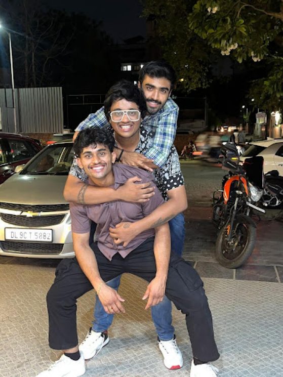 Three young men pose hugging each other smiling at the camera in front of a road with parked cars and a motorcylce