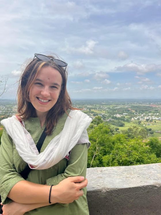 A young woman with arms crossed smiles directly at the camera with a green scenic background behind her