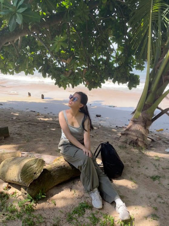 A young woman wearing sunglasses sits on a beach under a green tree