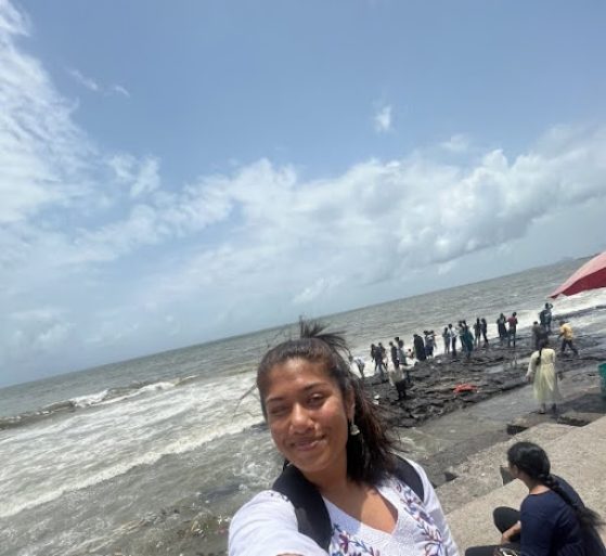 Individual standing by a large body of water, smiling, with dark hair pulled back