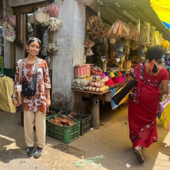  A smiling individual stands in front of a busy food market with various produce displayed on stands in the background