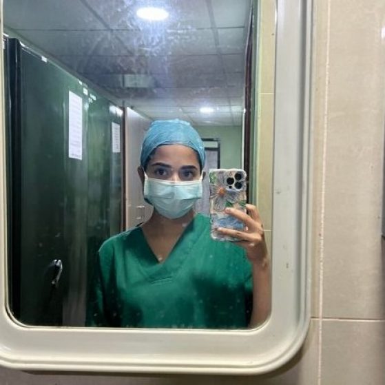 Varshini is taking a photo in the mirror wearing scrubs and a surgical face mask