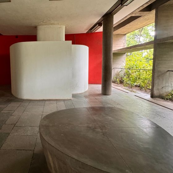 Tile floor with cement structures and a vibrant red wall. Greenery can be seen on the righthand side. 