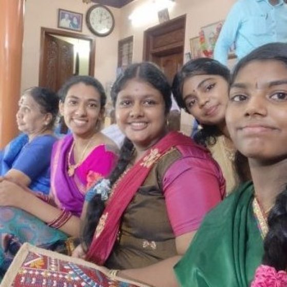 A group of people in colorful traditional attire sitting indoors, with a clock and plant visible in the background.