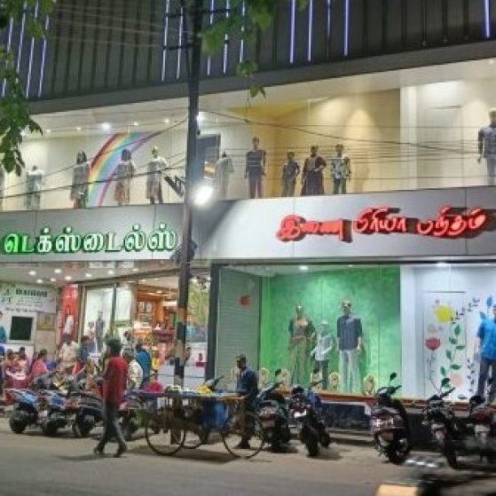 A bustling street scene in front of "Jaya Pa Textiles" with mannequins displayed above and people gathered below.
