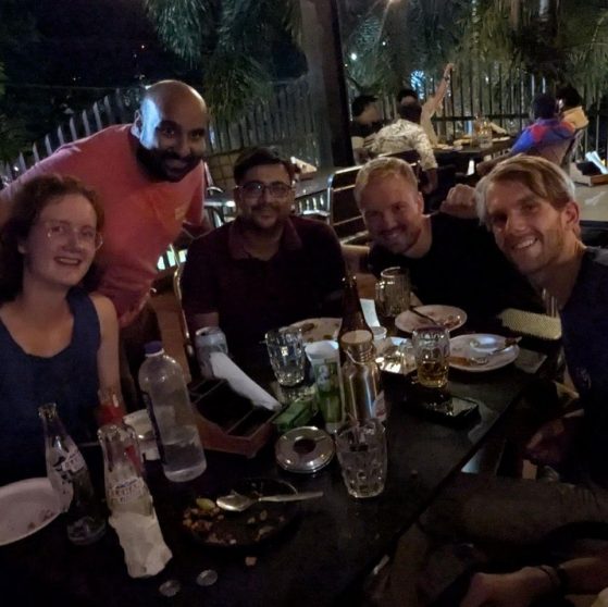 A group of six friends are sitting at a dinner table at night, smiling for a selfie