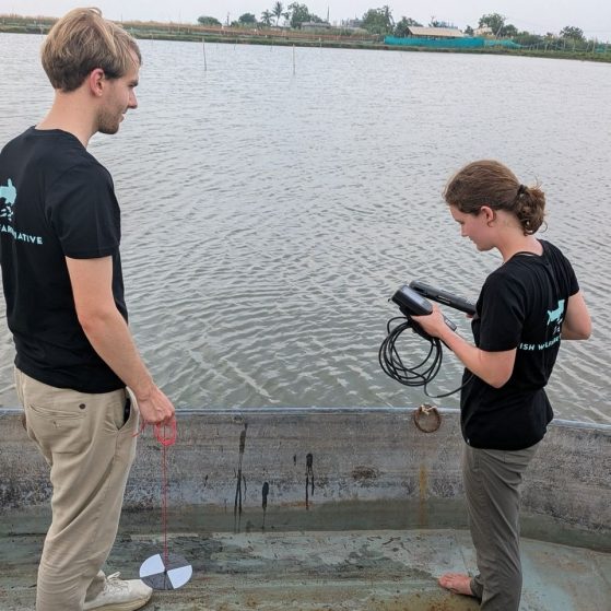 Annika and another individual, both wearing black shirts, are standing in a small aluminum boat on a body of water looking at a digital device