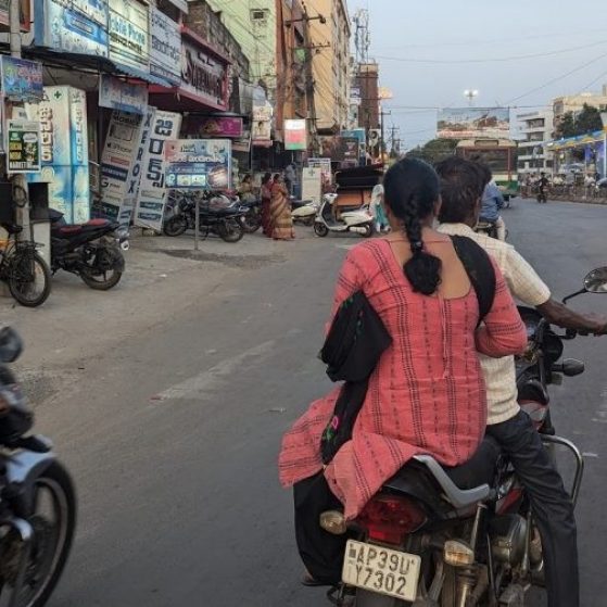 Two individuals ride on a motorcyle on a busy street in India 