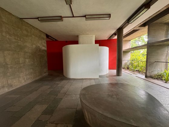 Tile floor with cement structures and a vibrant red wall. Greenery can be seen on the righthand side. 