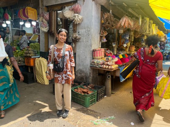  A smiling individual stands in front of a busy food market with various produce displayed on stands in the background