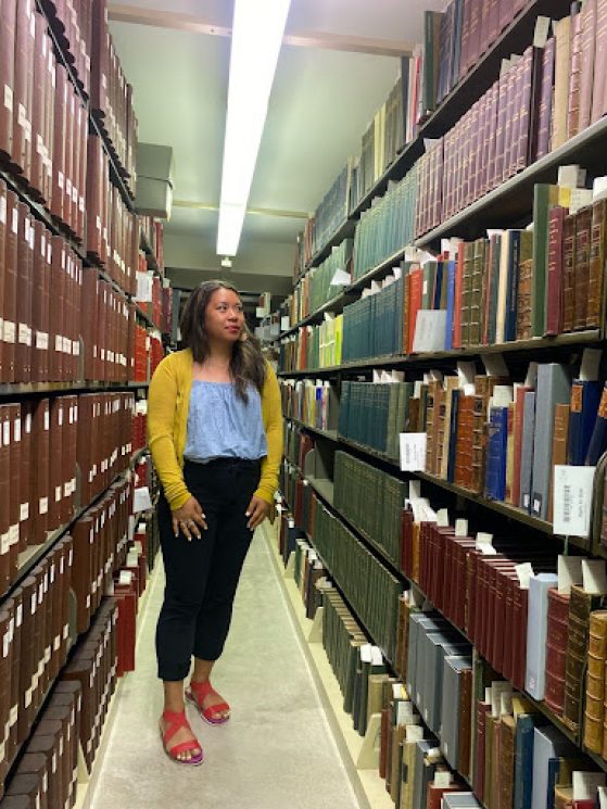 Janice Lobo Sapigao stands in the middle of two large floor to ceiling bookshelves filled with books. 