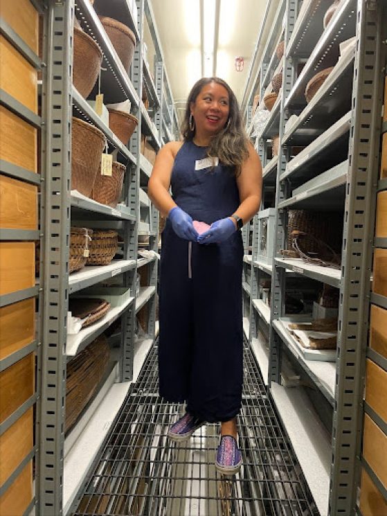 Janice Lobo Sapigao is wearing a navy dress with long hair standing in betweem floor to ceiling shelving units with boxes