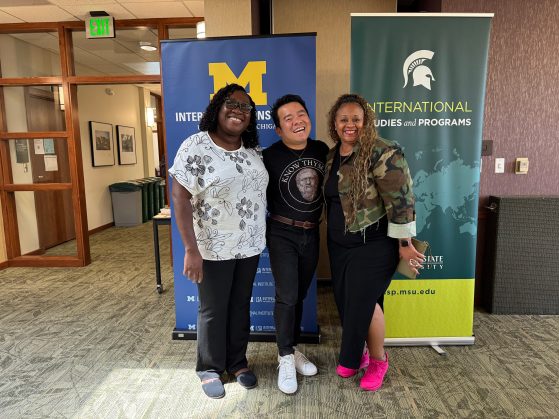 Three individuals stand side by side in front of U-M and MSU banners