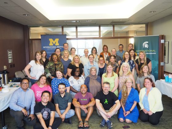 A large group photo of workshop participants sitting and standing in front of the International Institute and MSU banners smiling