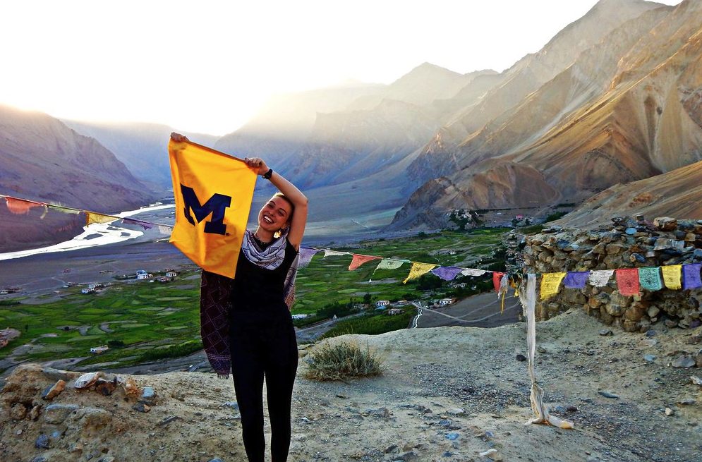 Female student holding a yellow M flag with peace flags behind her in a mountain setting
