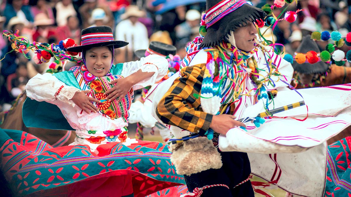 A colorful photo shows two people in traditional Peruvian costumes dancing at a festival. A woman in a red and teal patterned skirt and a man in a checkered jacket are in the foreground, with a crowd of people in the background.