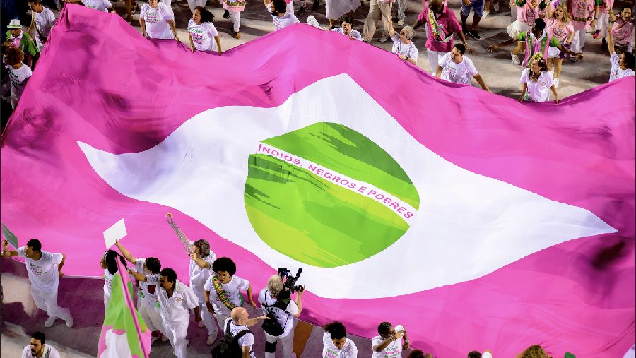 A large pink flag resembling the Brazilian flag, with a green circle and the words “Índios, Negros e Pobres,” held by people in a parade.