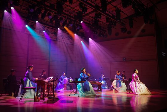 The King's Orchestra players are seated on a colorful stage playing a variety of musical instruments and wearing traditional Korean clothing