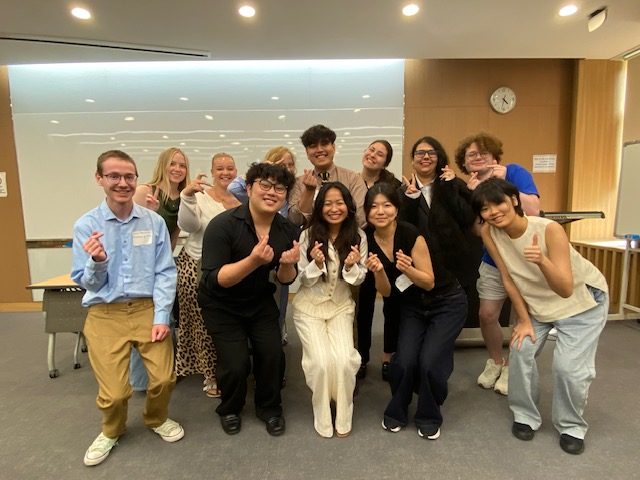 A group of students in business casual clothes pose for a picture while smiling and holding up their hands