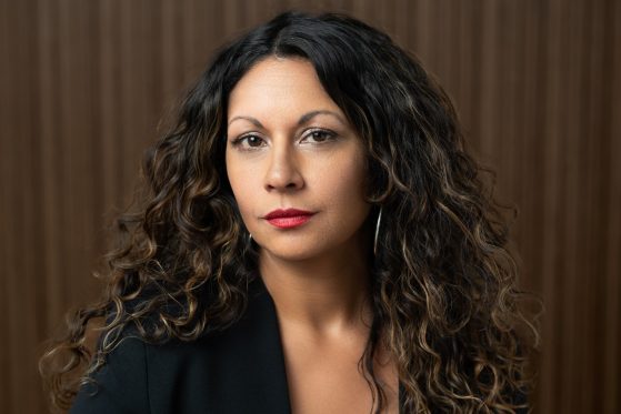 A woman with long, curly brown hair and red lipstick looks directly at the camera, wearing a black blazer, in front of a brown, vertically-striped background.
